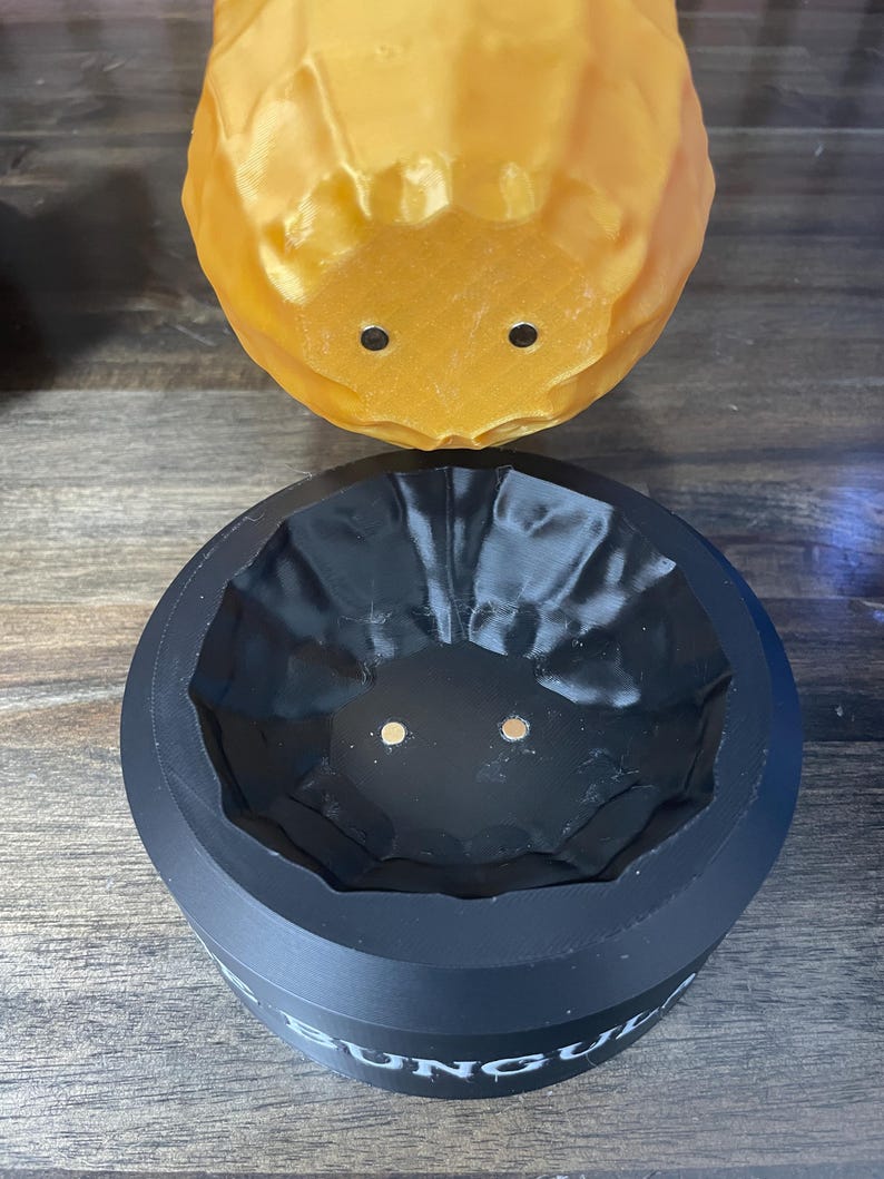 May include: A two-piece set featuring a yellow, textured, rounded object with two black dots, and a black, cylindrical container with the word "BUNG" visible. The container has a textured interior and two small gold dots.