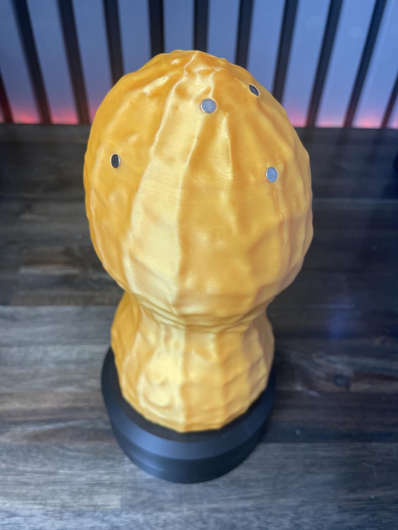 May include: A 3D-printed peanut-shaped object in a golden-yellow color, featuring a textured surface and several small, silver-colored circular accents. The peanut sits atop a black, cylindrical base, creating a unique decorative piece.
