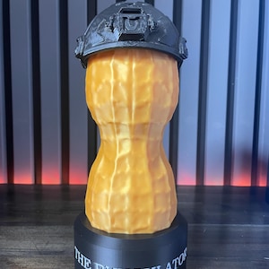May include: A 3D-printed sculpture of a peanut wearing a black tactical helmet. The peanut is a light orange color, with a textured surface. The base is black and has the text "THE BUNGULATOR" in white.