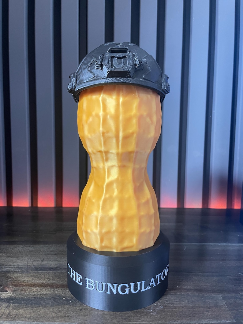 May include: A 3D-printed sculpture of a peanut wearing a black tactical helmet. The peanut is a light orange color, with a textured surface. The base is black and has the text "THE BUNGULATOR" in white.