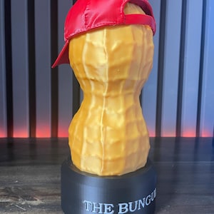 May include: A whimsical sculpture of a peanut wearing a red baseball cap, set atop a black base. The peanut is a golden yellow color, with the text "THE BUNGU" printed on the base. The cap is a vibrant red.