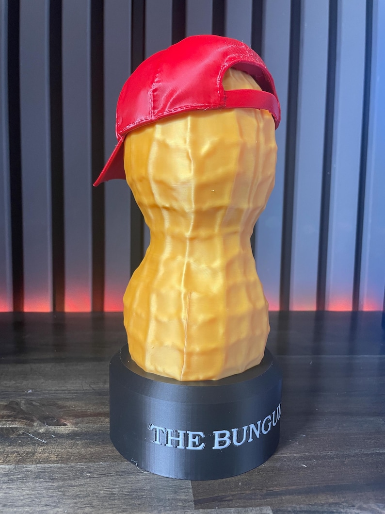 May include: A whimsical sculpture of a peanut wearing a red baseball cap, set atop a black base. The peanut is a golden yellow color, with the text "THE BUNGU" printed on the base. The cap is a vibrant red.