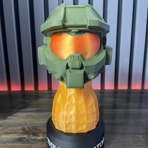 May include: A green and orange 3D-printed sculpture of a futuristic helmet, with an orange visor. The helmet sits atop a peanut-shaped base, which is mounted on a black circular base. The text "THE BUNGULATOR" is printed on the base.