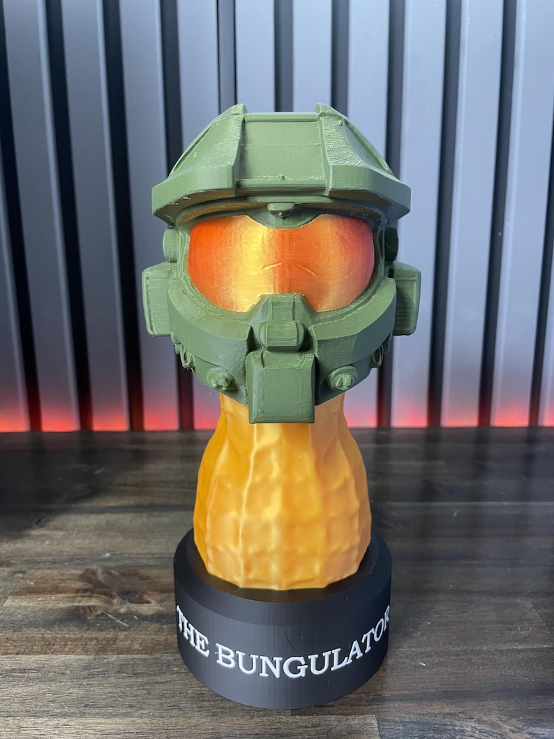 May include: A green and orange 3D-printed sculpture of a futuristic helmet, with an orange visor. The helmet sits atop a peanut-shaped base, which is mounted on a black circular base. The text "THE BUNGULATOR" is printed on the base.