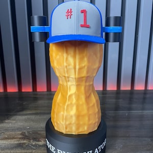 May include: A novelty sculpture of a peanut wearing a baseball cap with the number 1. The peanut is tan and rests on a black base inscribed with "THE BUNGULATOR". Two black cylinders are attached to the sides of the cap with blue bands.