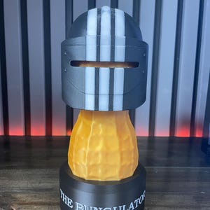 May include: A 3D-printed sculpture featuring a black and white striped helmet design atop a peanut-shaped body. The base is black with the text "THE BUNGULATOR" in white. The sculpture is a unique decorative item.