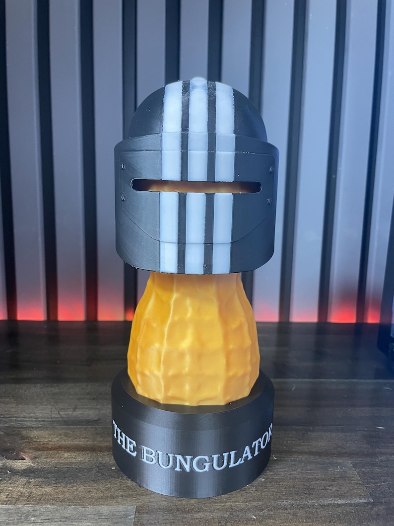 May include: A 3D-printed sculpture featuring a black and white striped helmet design atop a peanut-shaped body. The base is black with the text "THE BUNGULATOR" in white. The sculpture is a unique decorative item.