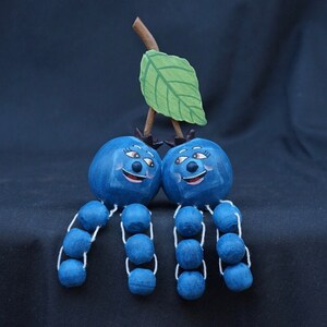 May include: Two blue blueberry figurines with painted faces and white stems. Each blueberry has six smaller blue blueberries attached to the stem. A green leaf is attached to the top of the stems.