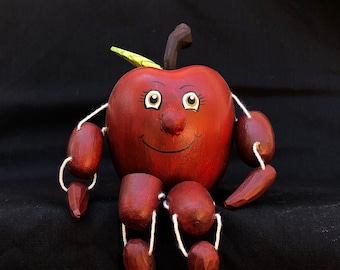 Puede incluir: Una figurita de manzana de madera roja con una cara pintada y una hoja verde en la parte superior. La manzana tiene una cara sonriente con ojos marrones y una nariz marrón. La manzana está sentada sobre una superficie negra con las piernas de madera extendidas.