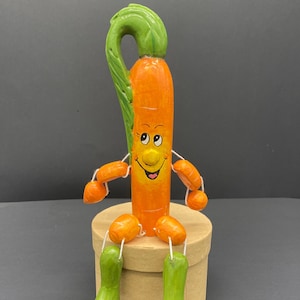 Op de afbeelding: Een handgeschilderd wortelfiguur met een glimlachend gezicht, dat groene laarzen draagt ​​en op een bruine doos zit. De wortel heeft een groen, bladerig topje.