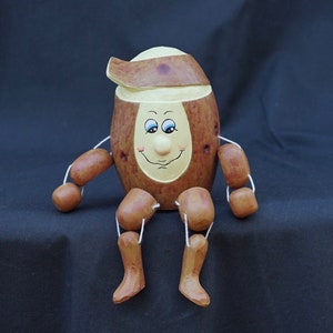 Może przedstawiać: Drewniana figurka ziemniaka z namalowaną twarzą i brązowym kapeluszem. Ziemniak ma brązowe i żółte malowane detale i siedzi ze skrzyżowanymi nogami.