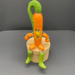 Op de afbeelding: Een figuur in de vorm van een wortel met een glimlachend gezicht en groen, bladerig haar zit op een bruine doos. De figuur heeft oranje, wortelvormige armen en benen.