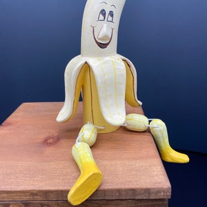 K&ouml;nnte beinhalten: Eine handbemalte Holzbananenfigur mit einem l&auml;chelnden Gesicht und beweglichen Gliedma&szlig;en. Die Banane ist gelb mit braunen Akzenten und sitzt auf einer Holzoberfl&auml;che.