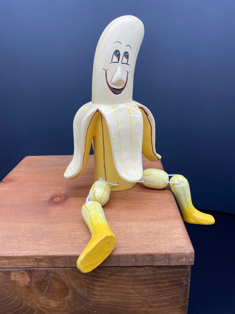 K&ouml;nnte beinhalten: Eine handbemalte Holzbananenfigur mit einem l&auml;chelnden Gesicht und beweglichen Gliedma&szlig;en. Die Banane ist gelb mit braunen Akzenten und sitzt auf einer Holzoberfl&auml;che.