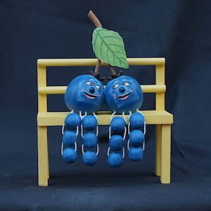 May include: Two blue blueberry figurines with painted faces sit on a yellow wooden bench. Each blueberry has a stem with a string of smaller blue blueberries hanging from it. A green leaf is attached to the top of the bench.