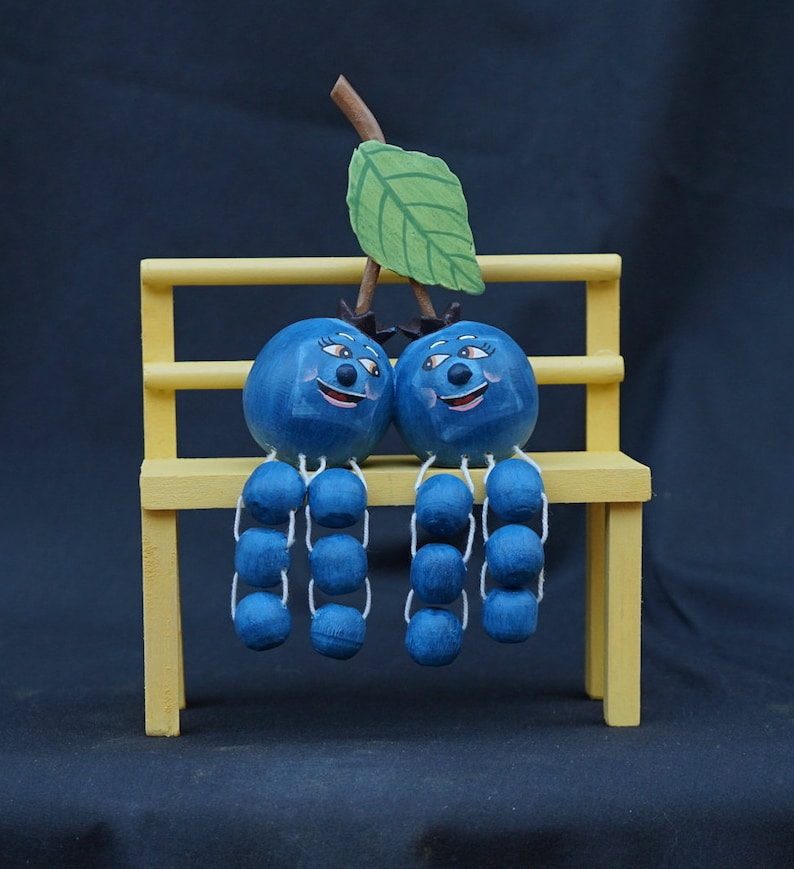 May include: Two blue blueberry figurines with painted faces sit on a yellow wooden bench. Each blueberry has a stem with a string of smaller blue blueberries hanging from it. A green leaf is attached to the top of the bench.