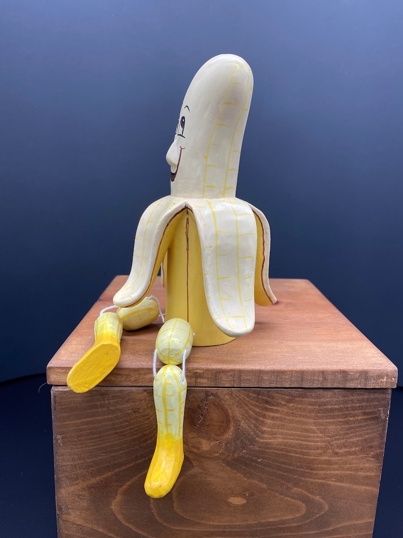K&ouml;nnte beinhalten: Eine Bananenfigur aus Holz mit einem l&auml;chelnden Gesicht und gelber und wei&szlig;er Farbe. Die Banane sitzt auf einer Holzkiste, wobei ihre Beine &uuml;ber den Rand h&auml;ngen.