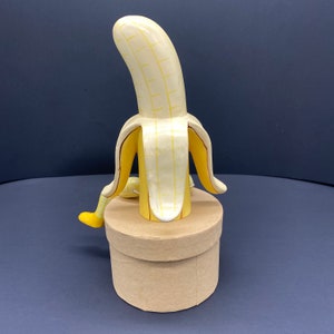 K&ouml;nnte beinhalten: Eine gelbe Bananen-Skulptur mit braunen Streifen, die aufrecht auf einem runden, braunen Sockel steht. Die Banane ist zur&uuml;ckgesch&auml;lt, um das Innere zu zeigen, das ebenfalls gelb ist.