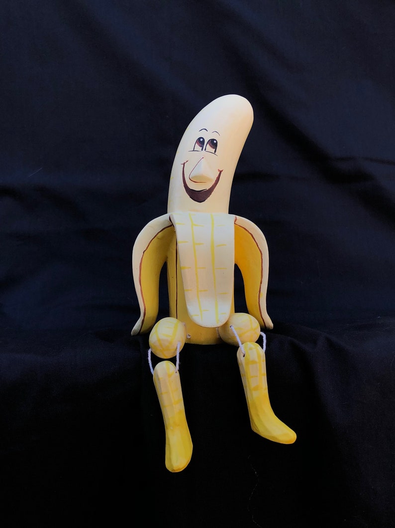 K&ouml;nnte beinhalten: Eine Holzpuppe in Form einer Banane mit einem l&auml;chelnden Gesicht, gelber und brauner Farbe und Gelenkbeinen.
