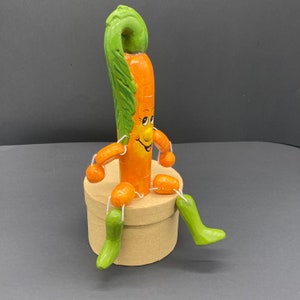 Op de afbeelding: Een grillig wortelfiguur met een glimlachend gezicht en een groene, bladerrijke bovenkant, zittend op een bruine kartonnen doos. De wortel heeft kleine wortelarmen en -benen.