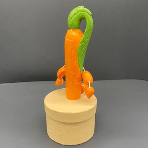Op de afbeelding: Een decoratief figuurtje in de vorm van een wortel met groene bladeren en kleinere oranje wortels die eraan hangen. Het figuurtje staat op een bruine kartonnen doos.