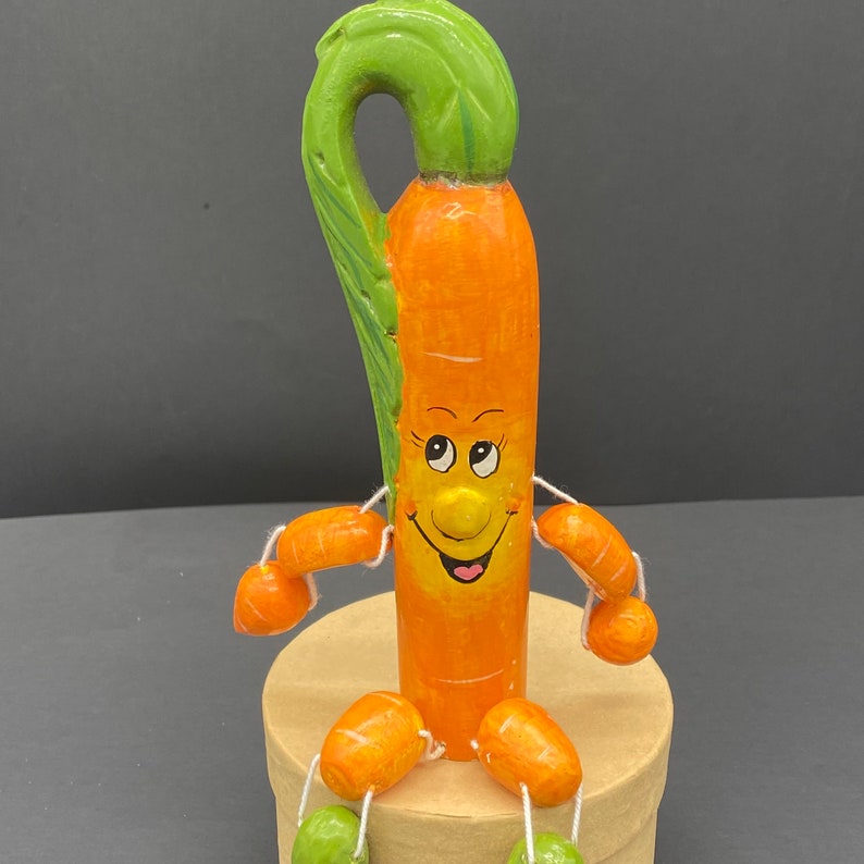 Op de afbeelding: Een figuur in de vorm van een wortel met een groen, bladerig bovendeel en een glimlachend gezicht. De wortel is oranje en heeft kleine, oranje wortelvormige armen en benen. Het figuur zit op een bruine basis.