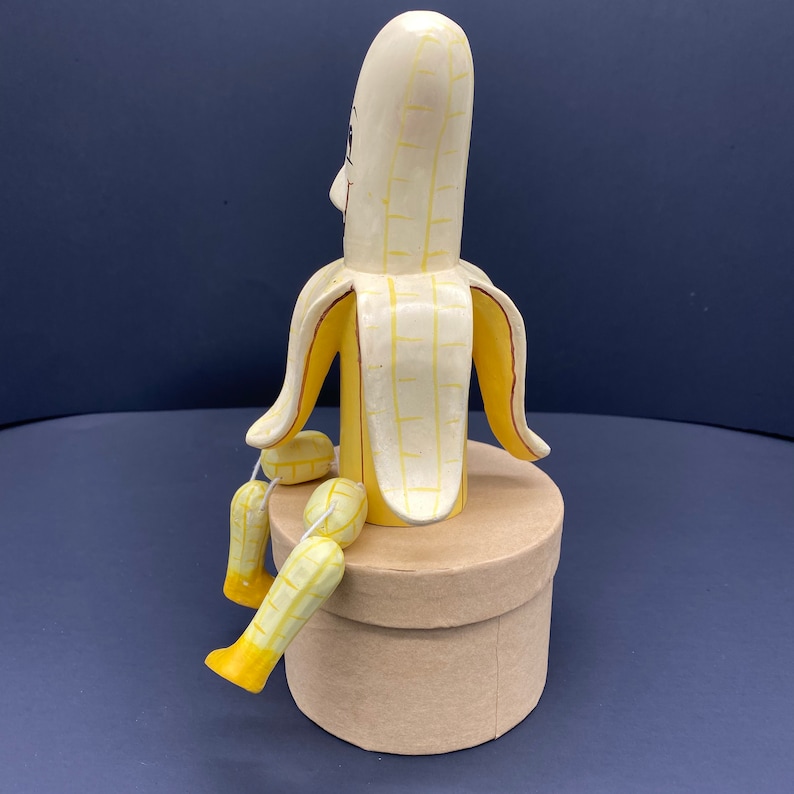 K&ouml;nnte beinhalten: Eine gelbe, bananenf&ouml;rmige Holzfigur mit einem Gesicht und Armen. Die Figur sitzt auf einem braunen Karton.