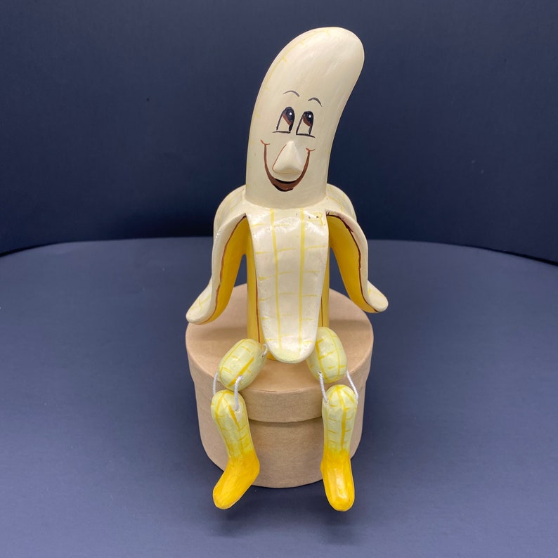 K&ouml;nnte beinhalten: Eine handbemalte Keramik-Bananenfigur mit einem l&auml;chelnden Gesicht und gelben und braunen Details. Die Banane sitzt auf einem braunen Sockel und hat ihre Beine in den Knien gebeugt.