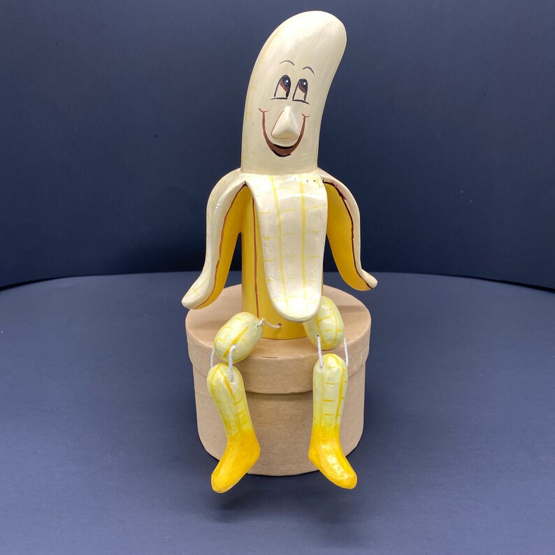 K&ouml;nnte beinhalten: Eine handbemalte Keramik-Bananenfigur mit einem l&auml;chelnden Gesicht und braunen Augen. Die Banane sitzt auf einem braunen Sockel und hat gelbe und braune Maldetails. Die Banane hat vier kleine bananenf&ouml;rmige Beine.
