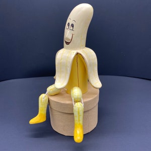 K&ouml;nnte beinhalten: Eine gelbe bananenf&ouml;rmige Figur mit einem l&auml;chelnden Gesicht und bemalten Details. Die Figur hat zwei Beine und sitzt auf einer braunen Box.