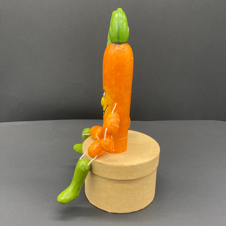 Op de afbeelding: Een handgeschilderd wortelfiguur met een glimlachend gezicht en een groene, bladerige bovenkant, zittend op een bruine kartonnen doos.