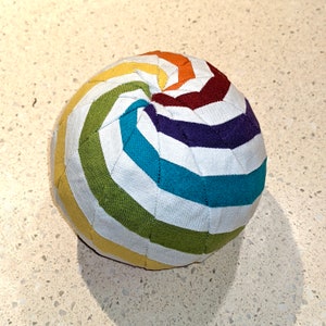 Puede incluir: Una bola de tela blanca con un patrón de espiral arcoíris. La bola está hecha de paneles de tela cosidos juntos, creando un efecto acolchado. Los colores de la espiral son rojo, naranja, amarillo, verde, azul, morado y blanco.