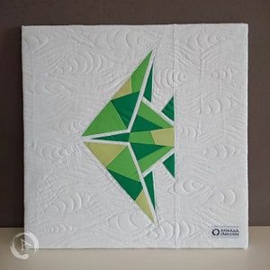 Könnte beinhalten: Eine weiße Leinwand mit einem geometrischen Fischdesign in Grün und Gelb. Der Fisch ist aus Stoff gefertigt und gesteppt. Die Leinwand hat einen weißen gesteppten Hintergrund mit einem Wellenmuster. Die Leinwand hat ein kleines Etikett in der rechten unteren Ecke, auf dem "Amarra Creaciones" steht.