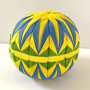 Puede incluir: Una bola de tela amarilla, azul y verde con un patrón geométrico. La bola está hecha de tela y está cosida en forma de esfera.