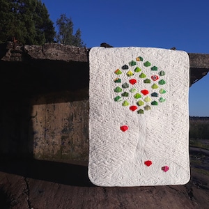Puede incluir: Una colcha blanca con un diseño de árbol verde y rojo. El árbol está hecho de cuadrados de tela y está rodeado de un fondo blanco. La colcha está colgada en una pared de hormigón.