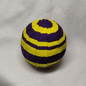 Puede incluir: Una esfera de tela rayada de morado y amarillo. La esfera está cosida en un patrón de rayas horizontales.