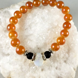 May include: A bracelet made with orange iridescent beads and black lava beads. The bracelet has a silver charm with a white opal stone in the centre.