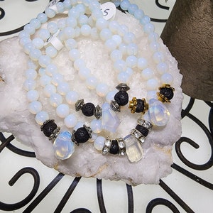 May include: Three white opal beaded bracelets with black lava beads and silver and gold accents. Each bracelet has a different charm, including a clear crystal, a teardrop-shaped opal, and a rectangular opal.