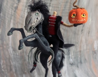 May include: A handcrafted Halloween-themed sculpture of the Headless Horseman riding a black horse. The rider wears a red jacket and black cape, holding an orange crocheted pumpkin. The horse stands on a wooden base.