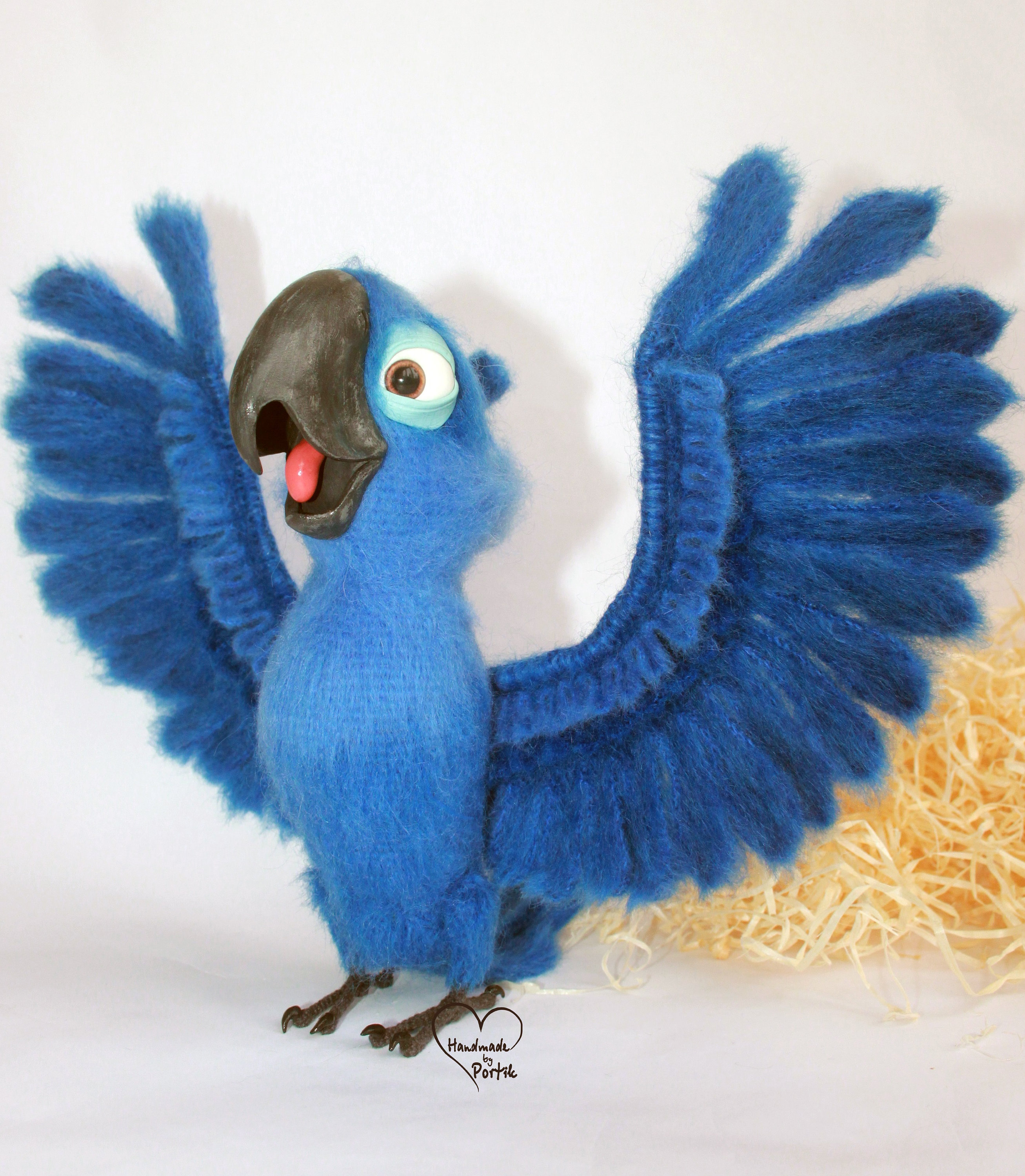 May include: A blue and black felt parrot with its wings spread wide. The parrot has a large beak and a bright blue body. The parrot is standing on a white surface with a brown background.