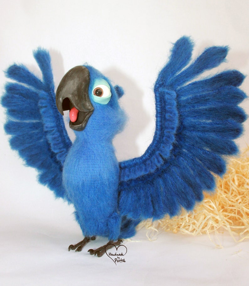May include: A blue and black felt parrot with its wings spread wide. The parrot has a large beak and a bright blue body. The parrot is standing on a white surface with a brown background.