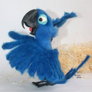 May include: Blue and black felt parrot toy with a red tongue and a white eye. The parrot is standing on a white surface with a brown background.