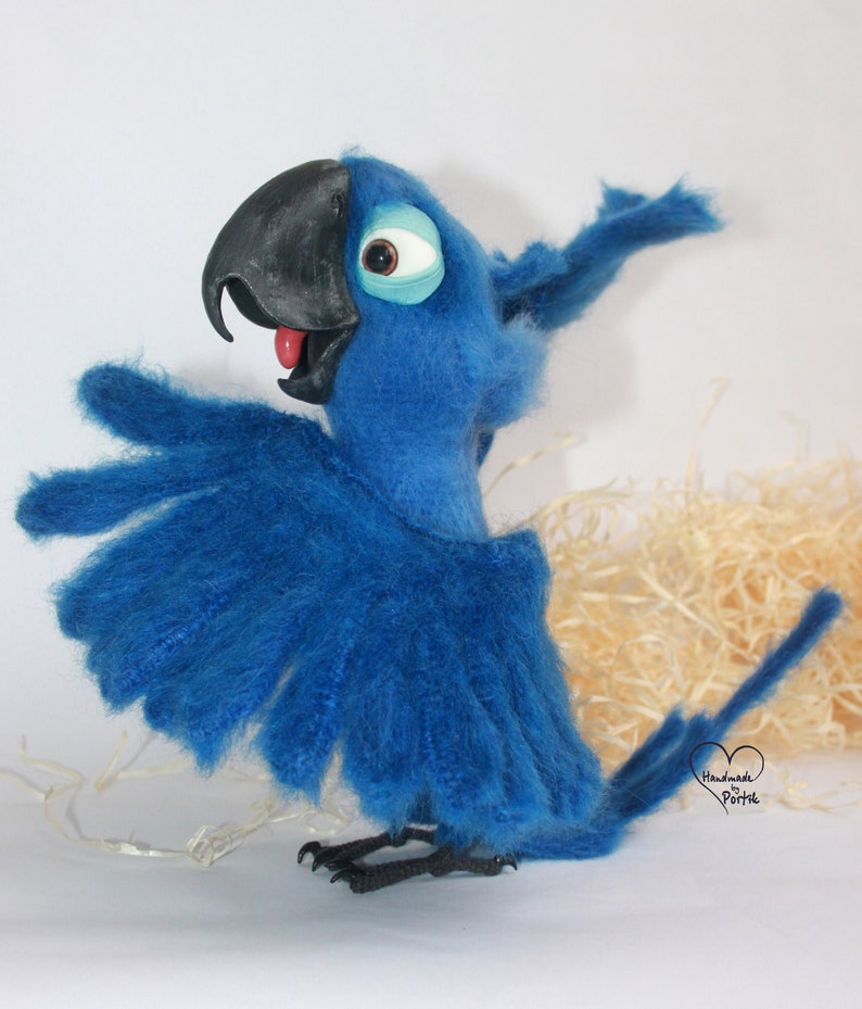 May include: Blue and black felt parrot toy with a red tongue and a white eye. The parrot is standing on a white surface with a brown background.