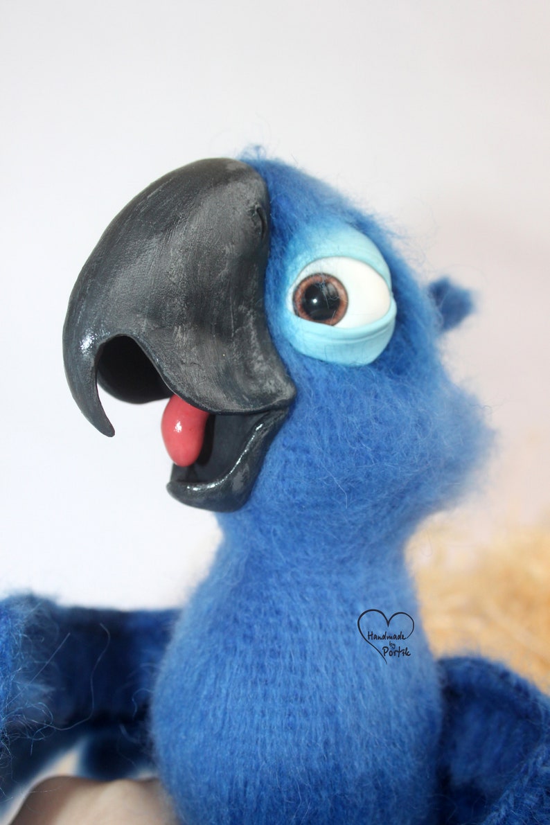May include: Blue and black felt parrot plush toy with a large, round eye. The parrot has a red tongue sticking out and is looking to the right. The toy is handmade and has a tag that says "Handmade by Portik".