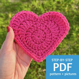 May include: A pink crocheted heart, held in a hand, with a blue banner that says "STEP-BY-STEP PDF pattern + pictures".