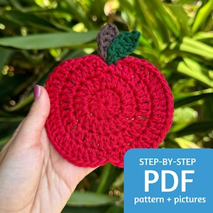 May include: A red crocheted apple with green leaves. The apple is being held in a hand. The text "STEP-BY-STEP PDF pattern + pictures" is in the bottom right corner of the image.