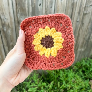 May include: A crocheted square with a yellow and brown sunflower design in the center. The square is made with a terracotta colored yarn.