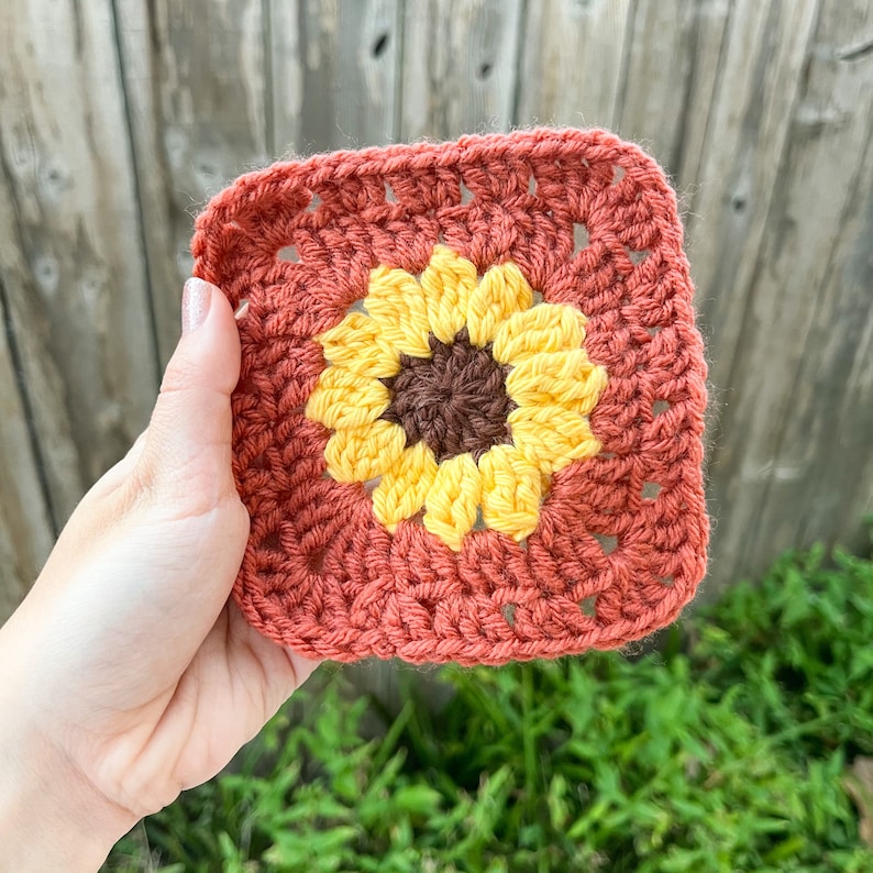 May include: A crocheted square with a yellow and brown sunflower design in the center. The square is made with a terracotta colored yarn.