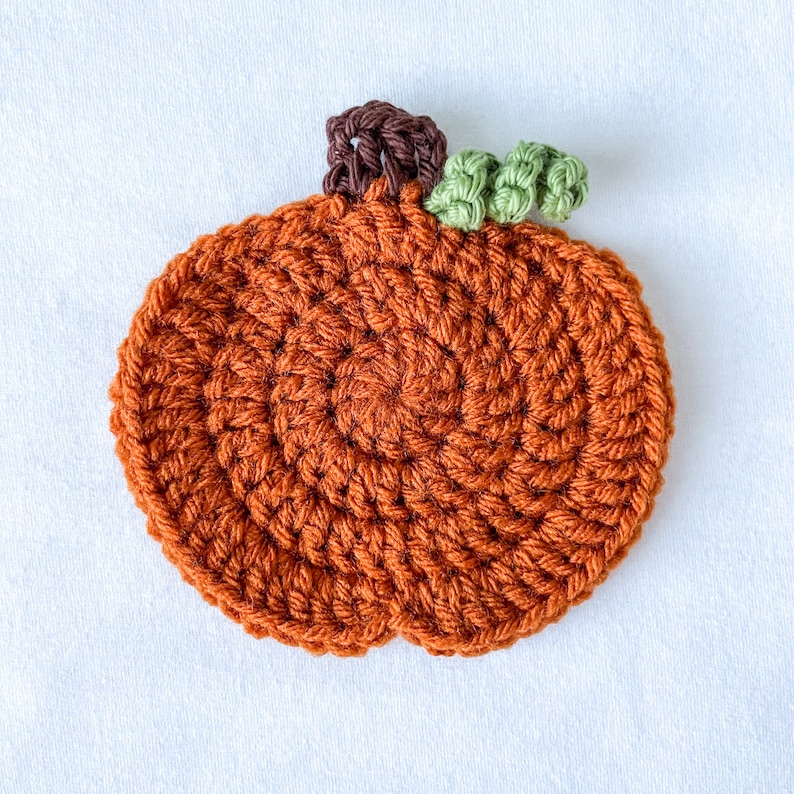 May include: A crocheted pumpkin with a brown stem and green leaves. The pumpkin is orange and has a spiral pattern.
