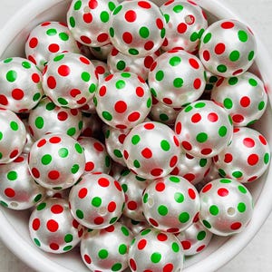 May include: A white bowl filled with pearlized beads. Each bead is decorated with red and green polka dots. The beads are round and have a glossy finish, creating a festive and playful appearance. The bowl is white and sits on a white surface.